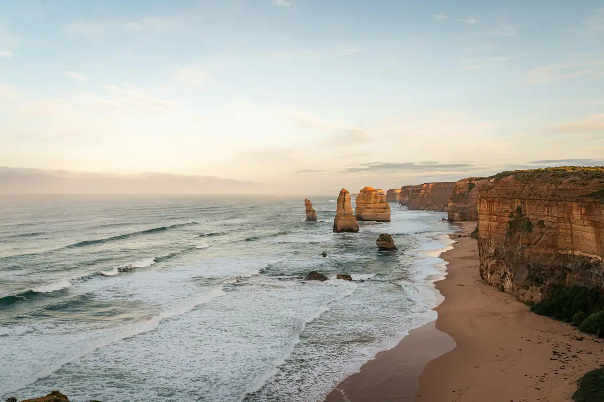 Soft sunrise light over the Twelve Apostles, with waves rolling onto the beach below the cliffs