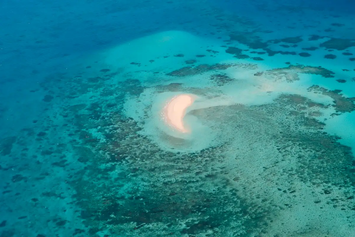 A small crescent sand cay sits among patchy coral reef in clear turquoise water on the Great Barrier Reef