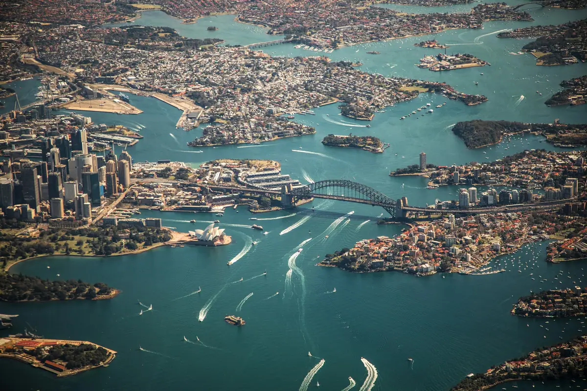 High aerial view of Sydney Harbour with the Harbour Bridge, Opera House, city towers and boat wakes across turquoise water