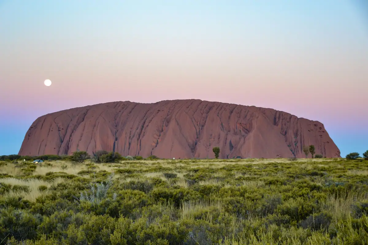 Uluru rises above green desert scrub under a pink and blue twilight sky with a full moon
