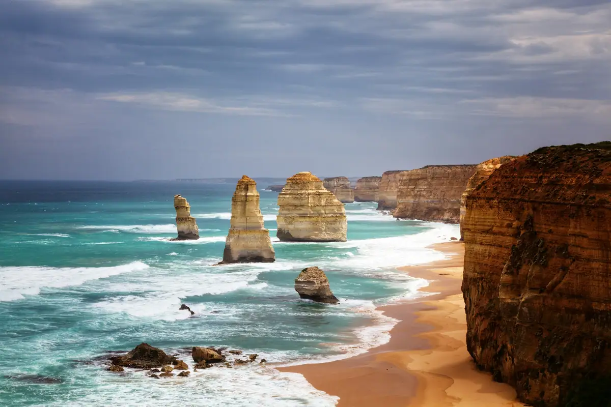 Limestone stacks of the Twelve Apostles rising from white surf beside towering golden cliffs under a moody sky