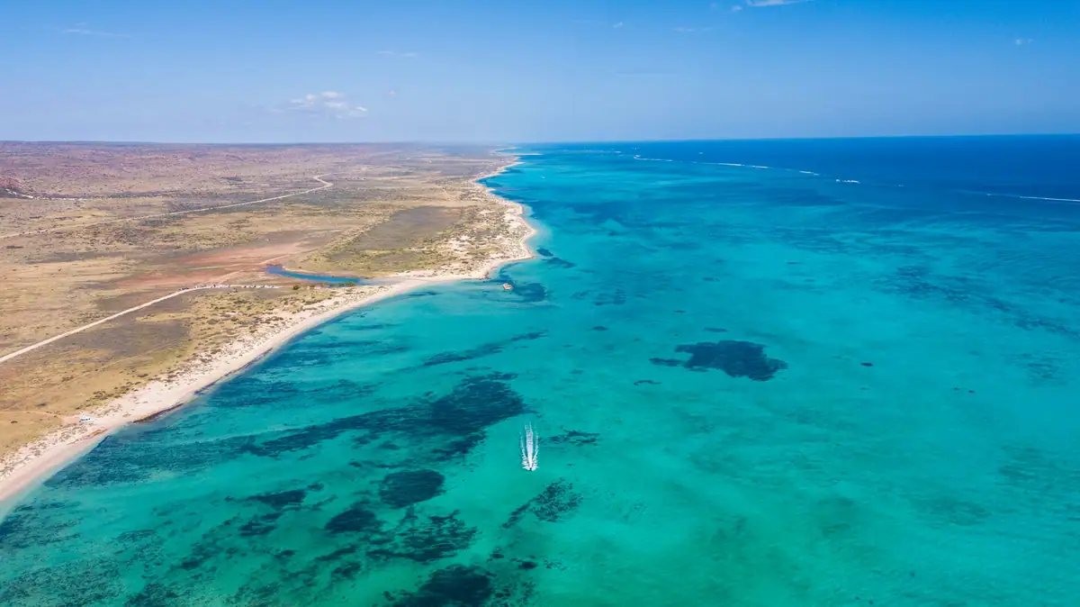Aerial view of Ningaloo's turquoise lagoon and reef beside a pale sandy coastline, with a small boat on the water