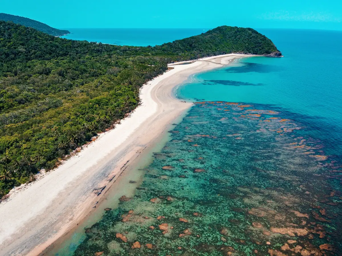 Aerial view of rainforest running down to a pale curving beach and fringing reef at Cape Tribulation, with turquoise sea beyond