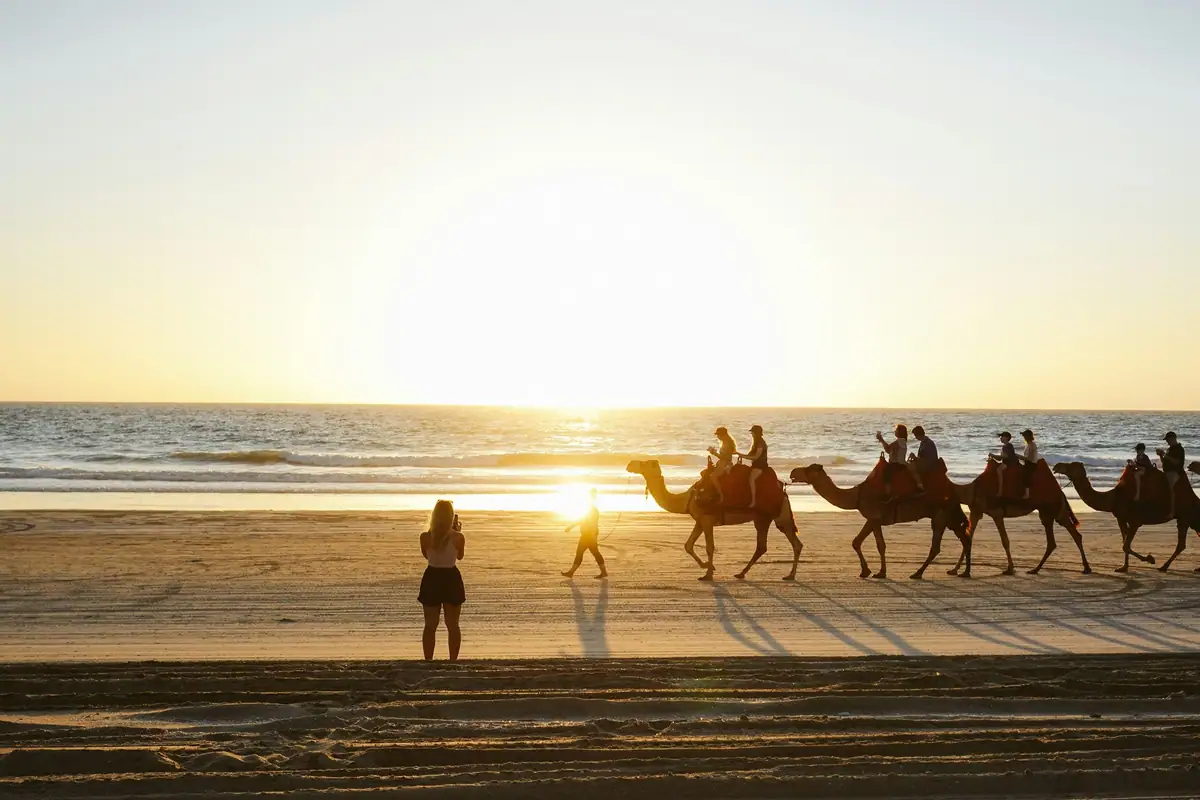 A camel train with riders walks along the beach at Broome into the setting sun as a person watches from the sand