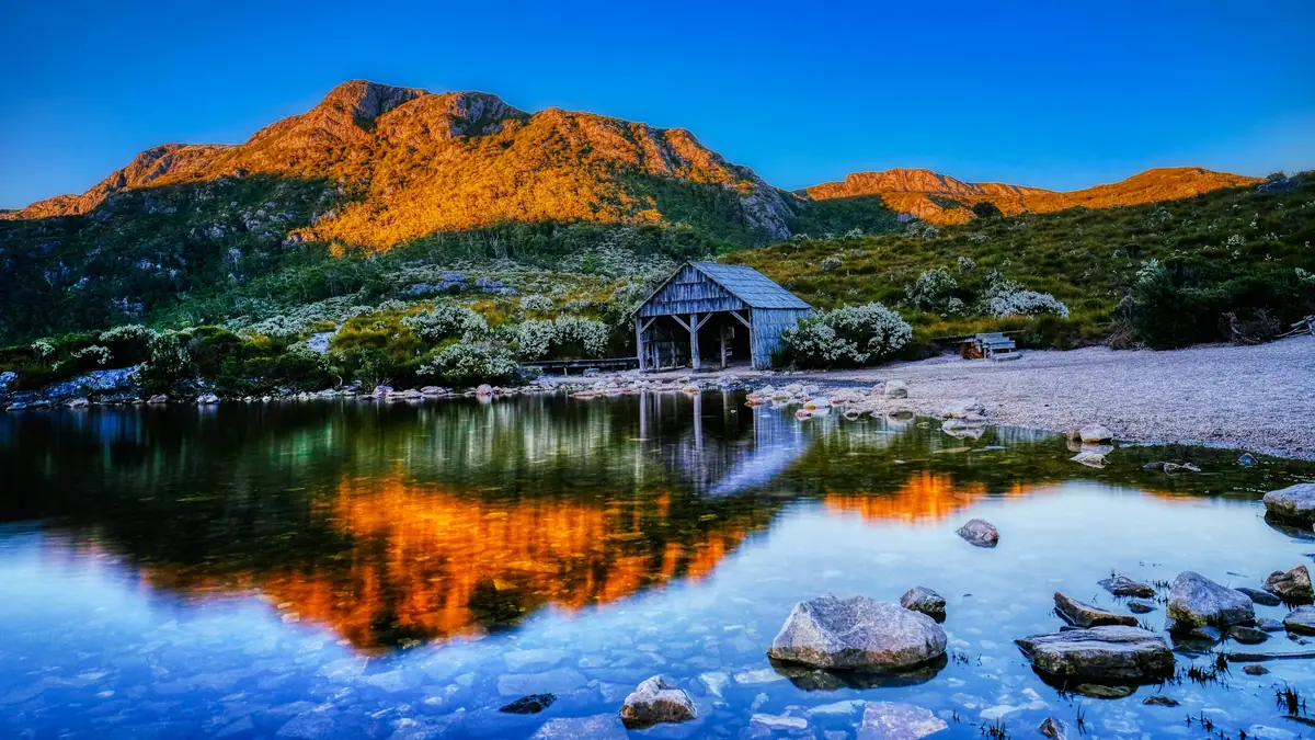 Cradle Mountain glowing orange at sunrise above the Dove Lake boatshed, its peaks mirrored in the still lake