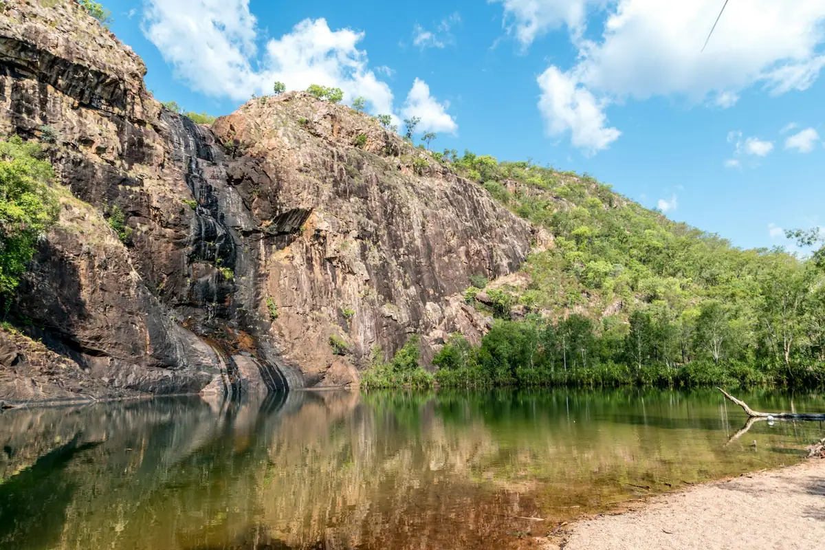 A calm plunge pool with a sandy edge reflects a streaked sandstone escarpment and green woodland in Kakadu