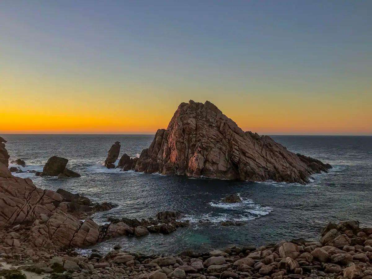 Sugarloaf Rock silhouetted against an orange sunset sky, with waves washing over the granite shore near Cape Naturaliste