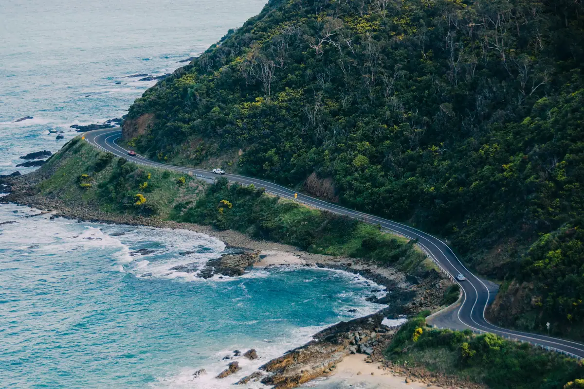 Aerial view of the Great Ocean Road winding along the coast between forested hills and rocky surf